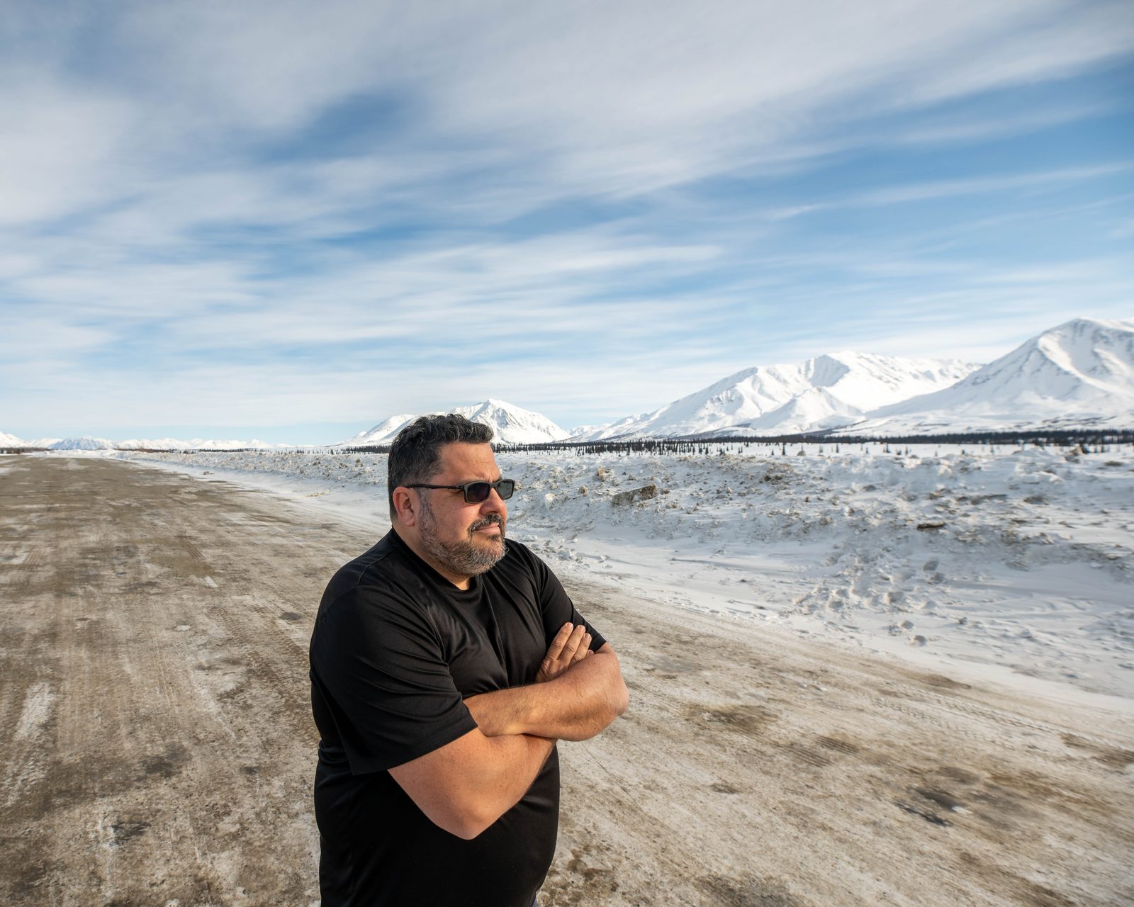 Juan in sunglasses and a T-shirt on a snowy road, mountains behind him